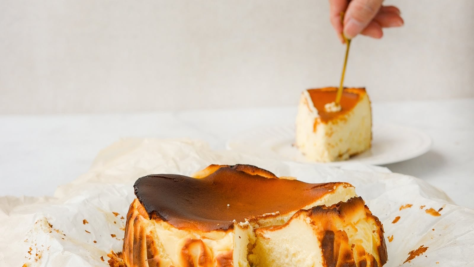 Basque burnt cheesecake utuh berbalut kertas baking dengan permukaan gosong karamel, satu potong terangkat