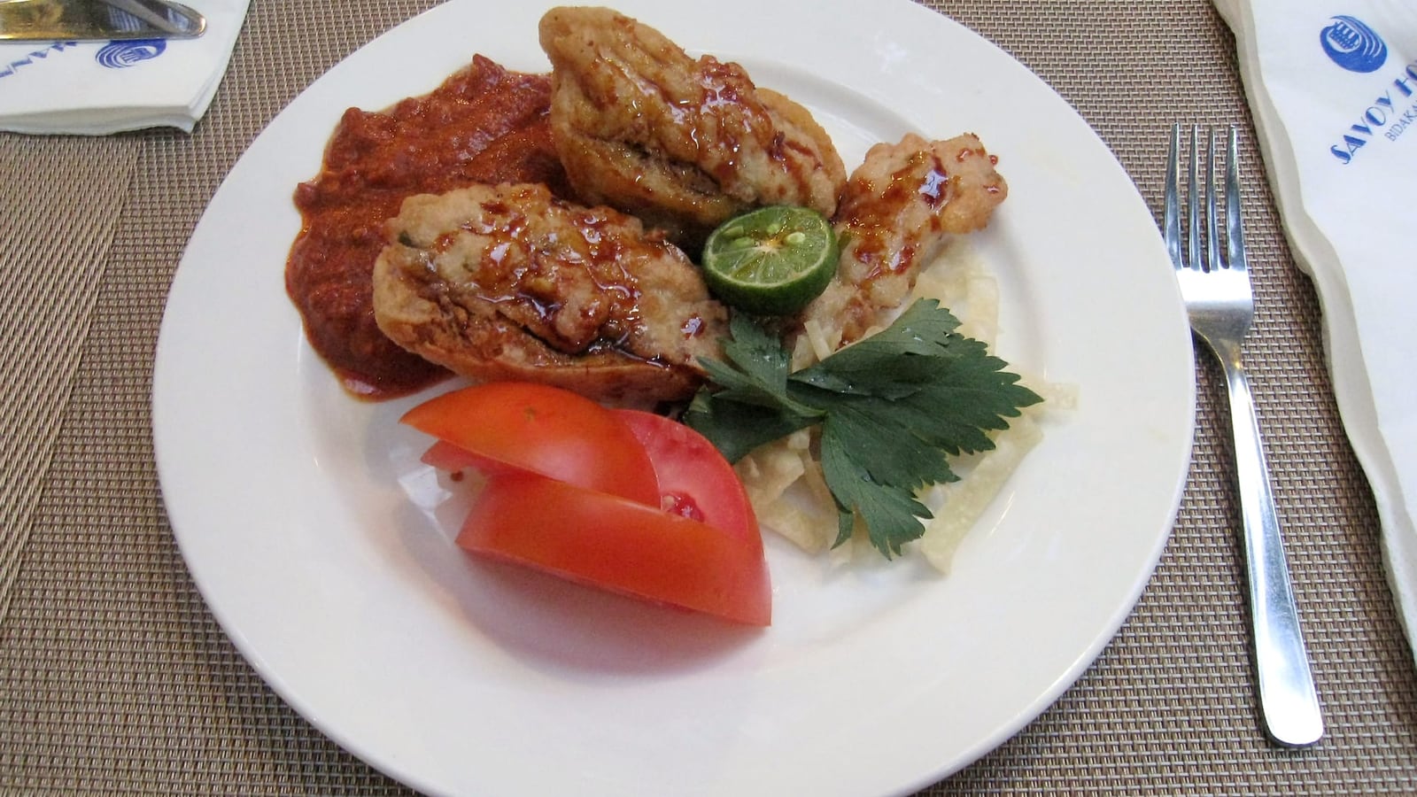 Batagor Bandung goreng keemasan disiram saus kacang, dengan jeruk limau dan irisan tomat di piring putih