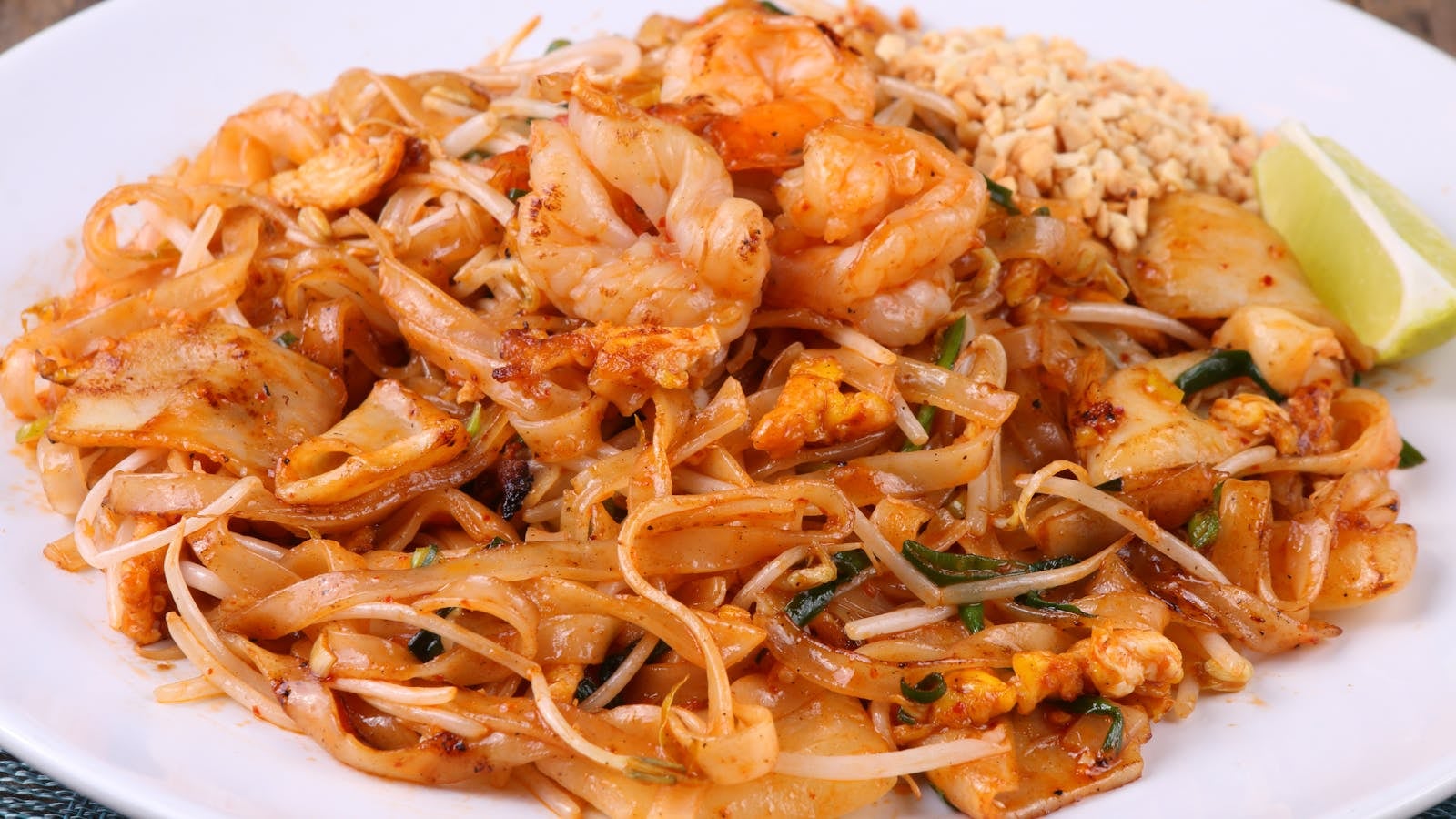 Pad thai udang besar dengan kacang tanah, tauge, jeruk nipis, dan daun bawang di piring putih